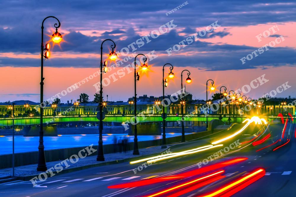 На фотографии изображён Троицкий мост, ярко освещённый в вечернее время. Закатное небо отражается в водах реки Невы, создавая живописную и спокойную атмосферу. Свет фонарей и световые следы машин подчёркивают динамику города. Троицкий мост – один из знаковых мостов Санкт-Петербурга, соединяющий берега Невы.